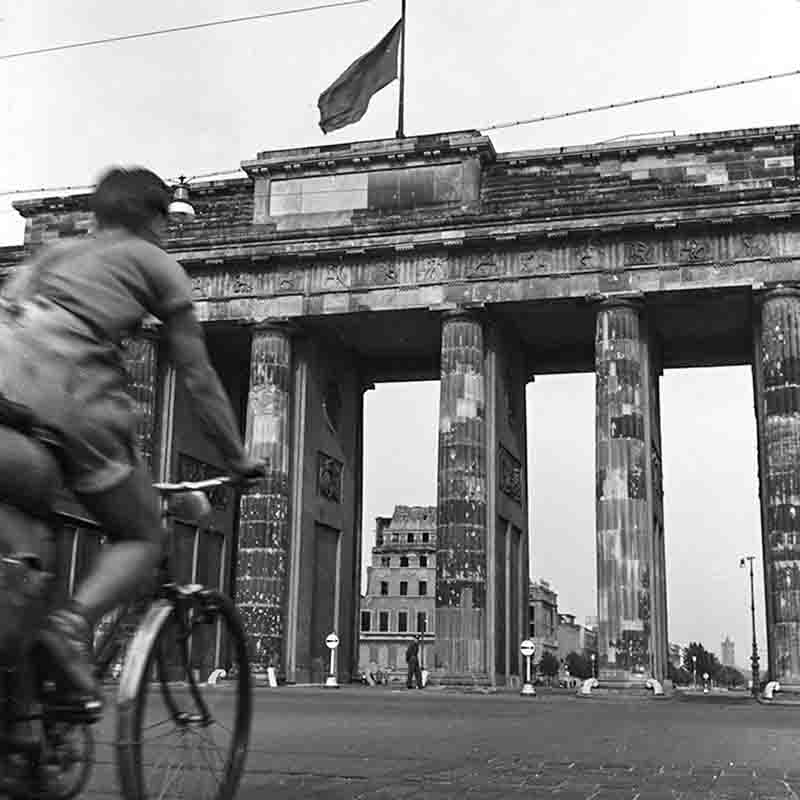Eine Schwarz-Weiß-Aufnahme des Brandenburger Tors in Berlin, aufgenommen aus einer niedrigen Perspektive. Im Vordergrund fährt ein Radfahrer, dessen Bewegung verschwommen ist.