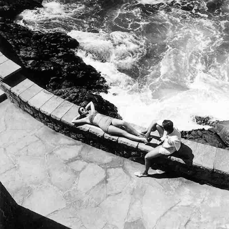 Eine Schwarz-Weiß-Aufnahme aus der Vogelperspektive, die zwei Personen an einer steinernen Ufermauer in Cabo Blanco zeigt. Eine Frau liegt auf dem Rücken und sonnt sich auf der Mauer, die Hände hinter dem Kopf verschränkt, während ein Mann in kurzen Hosen neben ihr sitzt.