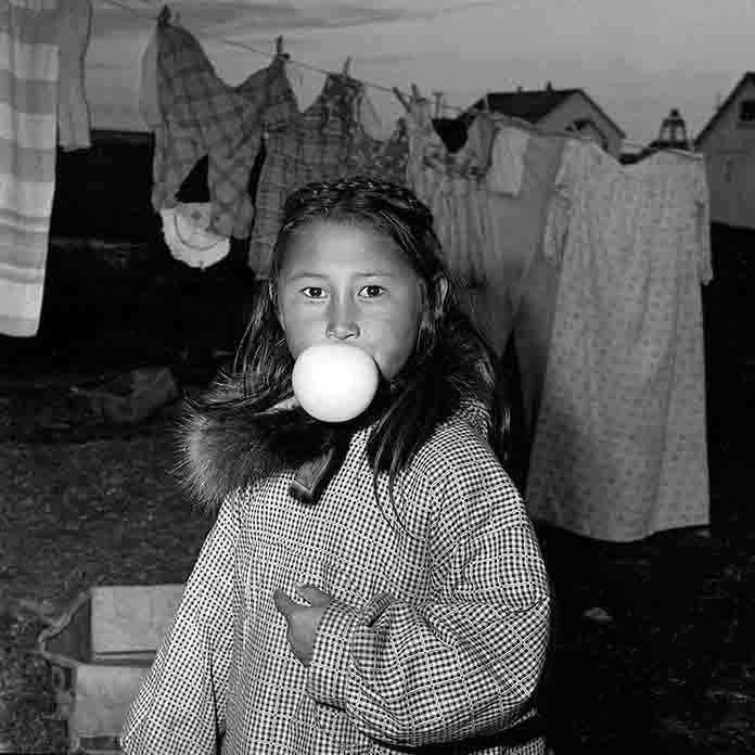 Eine Schwarz-Weiß-Nahaufnahme eines Inuit Mädchens, das einen großen Kaugummiball bläst, der fast ihren Mund und ihre Nase bedeckt. Sie trägt einen karierten Parka mit Pelzbesatz an der Kapuze.