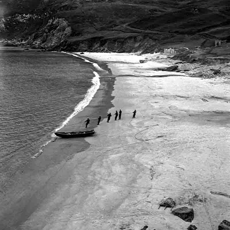 Eine Schwarz-Weiß-Luftaufnahme oder erhöhte Perspektive eines sandigen Strandes an einer felsigen Küste. Fünf oder sechs Personen ziehen an einem Seil, um ein kleines Boot, das von den Wellen umspült wird, vom Wasser auf den Strand zu ziehen.