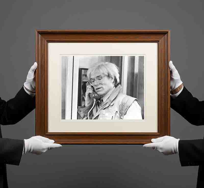 Schwarz-Weiß-Fotografie im braunen Holzrahmen mit weißem Passepartout, gehalten von zwei Händen in weißen Handschuhen. Das Foto zeigt den Künstler Andy Warhol in Nahaufnahme, der ein Telefongespräch führt und dabei durch die Verglasung oder Struktur einer Telefonzelle blickt.