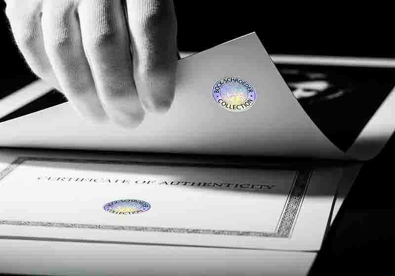 Weiße behandschuhte Hand hält ein Blatt Papier mit einem holografischen Echtheitssiegel, das einen Teil eines 'Certificate of Authenticity',Echtheitszertifikat freigibt.