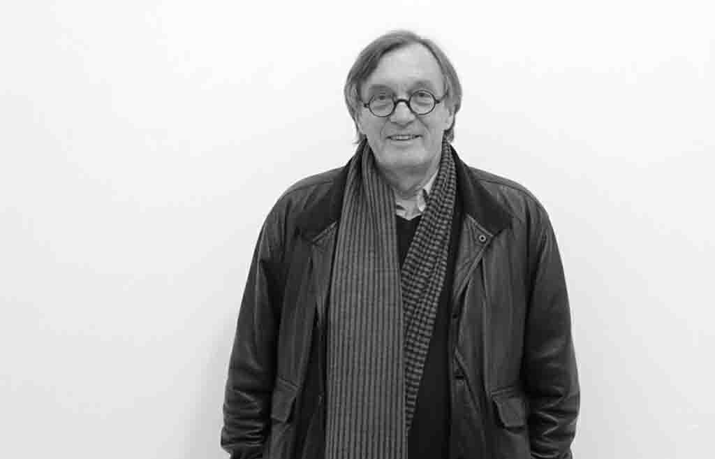 Ein Porträtfoto des Schweizer Künstlers Edy Brunner, der eine schwarze Lederjacke, einen dunkelblauen Pullover und einen grauen Schal trägt, vor einem weißen Hintergrund.