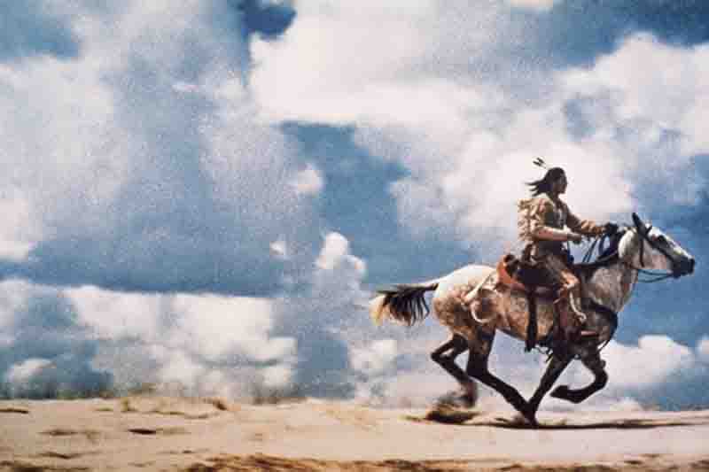 Ein Reiter mit Feder im Haar galoppiert auf einem weißen Pferd durch eine karge, sandige Landschaft unter einem dramatisch bewölkten Himmel.