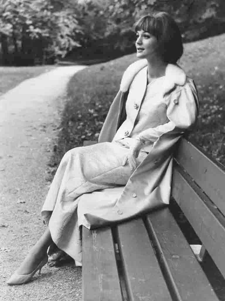 Ein Model in einem hellen Kostüm und einem über die Schultern gelegten Mantel mit Pelzkragen entspannt auf einer Parkbank.