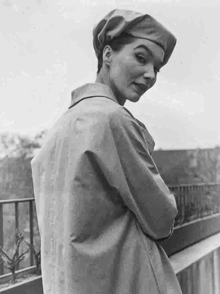 Porträt eines Models in einem hellen Mantel und einer voluminösen Baskenmütze. Der Schulterblick und der präzise Lidstrich betonen die zeitlose Eleganz. Die dezente Knickfalte im Foto unterstreicht den authentischen Vintage-Charakter dieser klassischen Außenaufnahme.